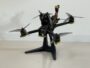 Renn-Quadcopter mit schwarzem Carbonrahmen und vier Propellern, gelben Motorabdeckungen und nach vorn gerichteter Kamera, auf einem schwarzen Ständer vor einer weißen Wand stehend; oben sichtbarer XT60-Stecker und Kabel.