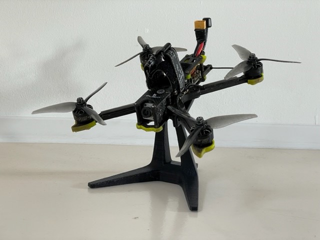 Renn-Quadcopter mit schwarzem Carbonrahmen und vier Propellern, gelben Motorabdeckungen und nach vorn gerichteter Kamera, auf einem schwarzen Ständer vor einer weißen Wand stehend; oben sichtbarer XT60-Stecker und Kabel.
