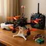 FPV-Quadrocopter mit vier leuchtend orangefarbenen Propellern und erhöhter Antenne steht auf einem weißen 3D-gedruckten Tischständer auf einem Holztisch; im Hintergrund Fernsteuerung, FPV-Brille und zwei grüne Akkus zu sehen.