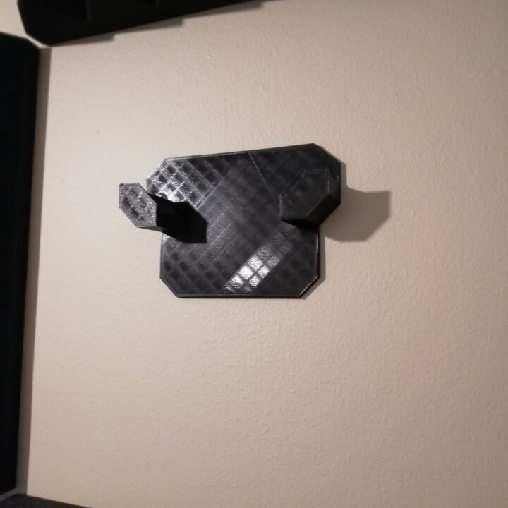 Schwarzes 3D-gedrucktes Wandhalterungsstück mit zwei abgewinkelten Haken und sichtbarem Schichtraster, montiert an einer hellen Wand.