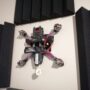 FPV-Rennquadrocopter mit Carbonrahmen, vier sichtbaren Motoren und rosa umwickelten Armen sowie einer kleinen gelben Antenne, an einer hellen Wand zwischen schwarzen Akustikschaumplatten montiert.