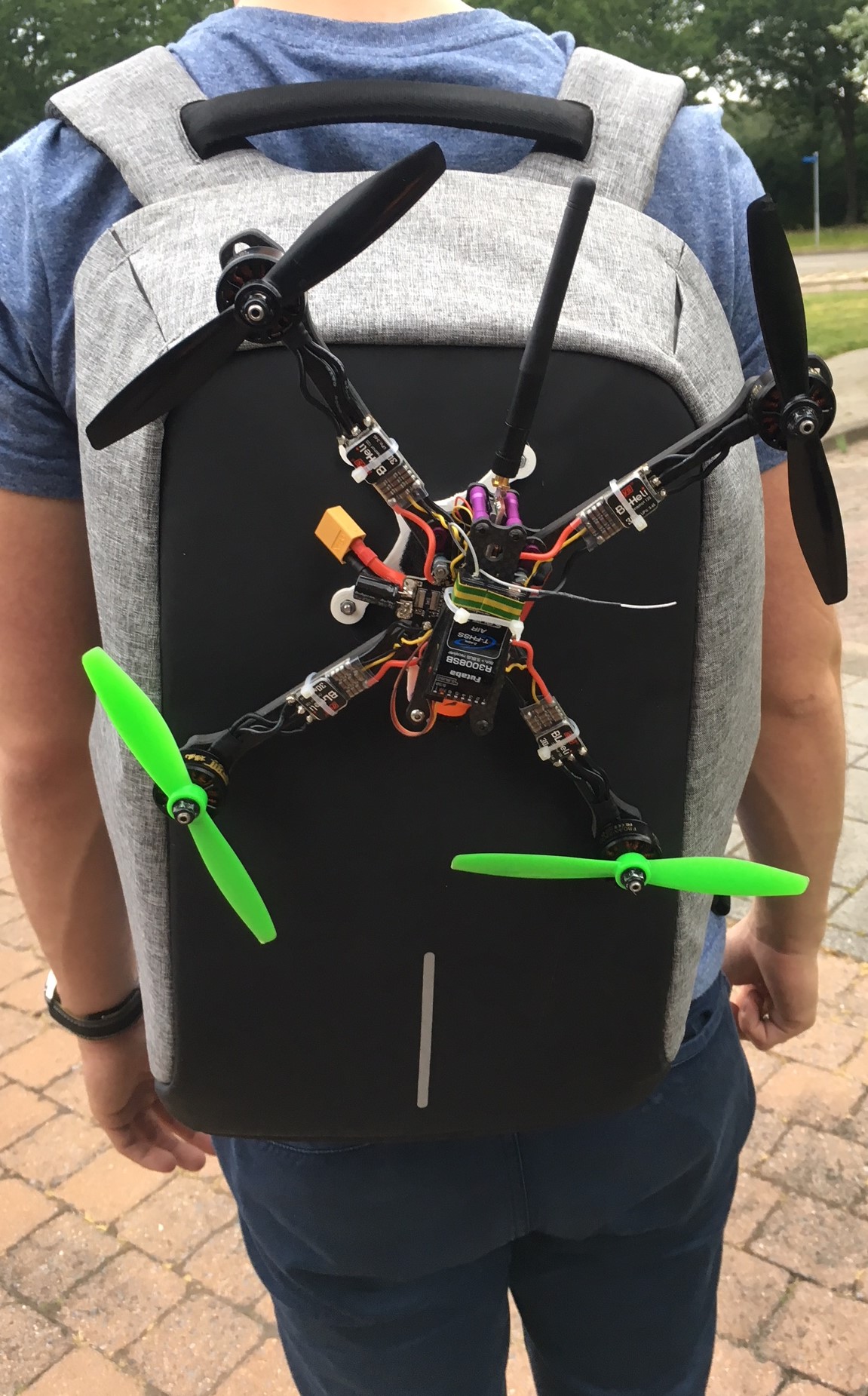 Person von hinten mit grauem Rucksack, an dem ein offener Racing-Quadrocopter mit schwarzem Rahmen, sichtbaren Kabeln, Antenne und zwei grünen sowie zwei schwarzen Propellern befestigt ist.