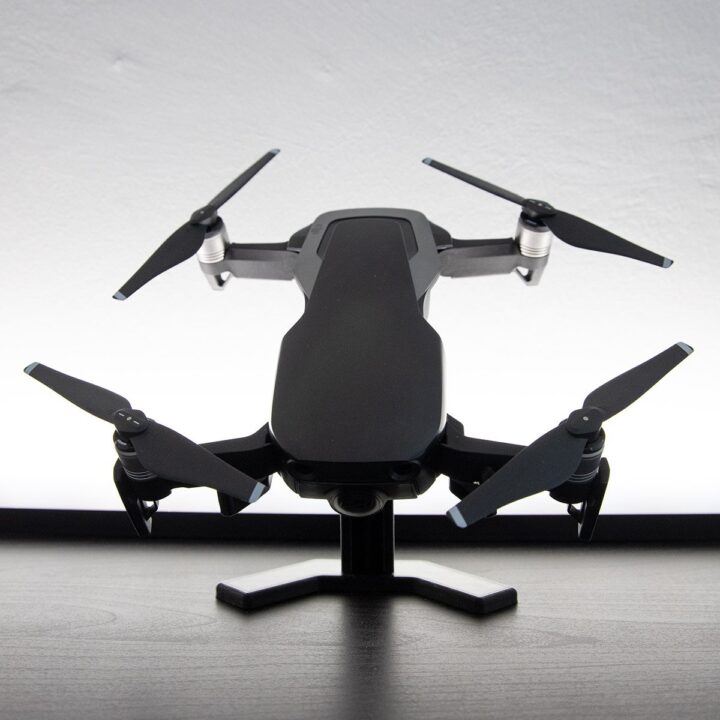 dronestand1 Schwarze Vierrotor‑Drohne mit ausgeklappten Propellern auf einem kleinen Ständer, frontal auf einem Tisch vor hellem Hintergrund.