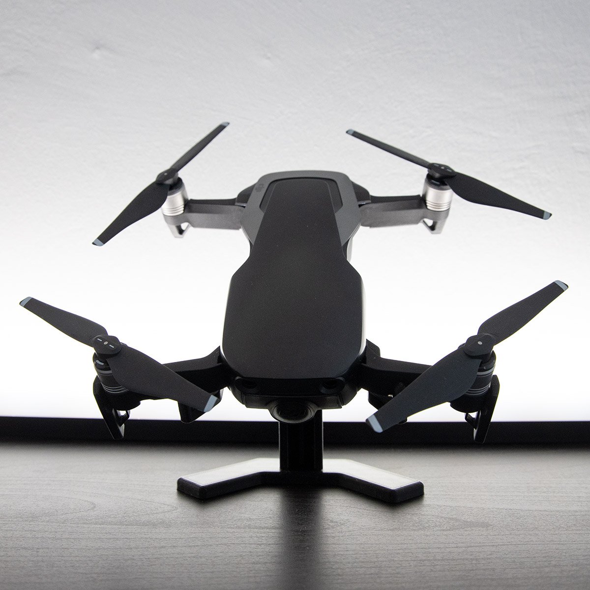 dronestand1 Schwarze Vierrotor‑Drohne mit ausgeklappten Propellern auf einem kleinen Ständer, frontal auf einem Tisch vor hellem Hintergrund.
