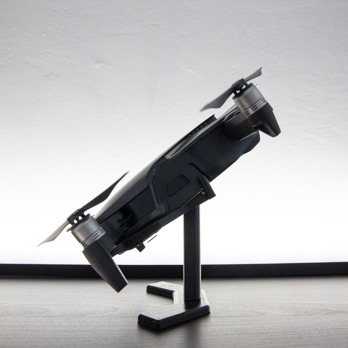 Kompakter schwarzer Quadrocopter auf einem schwarzen Ständer, schräg nach oben geneigt mit sichtbaren Propellern vor hellem, gleichmäßig beleuchtetem Hintergrund.