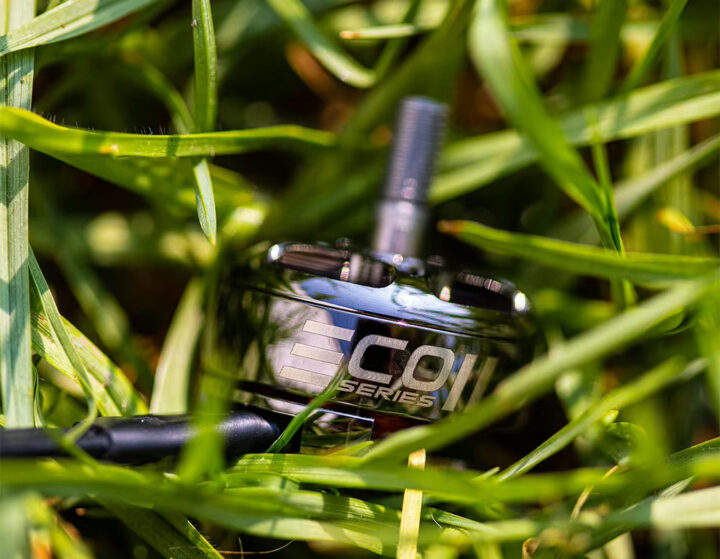 FPV-Drohnenmotor mit glänzendem Metallgehäuse, sichtbarer Serienbeschriftung und hervorstehender Antriebswelle, liegend zwischen dichtem grünem Gras.