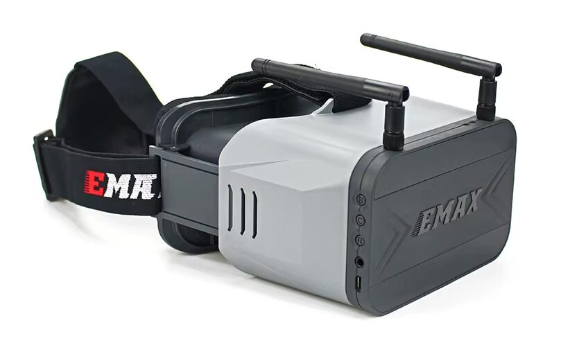 Graue und schwarze FPV-Drohnenbrille mit aufgesetzten Antennen, seitlichen Bedienknöpfen, auffälligem EMAX-Logo und verstellbarem Kopfband, freigestellt vor weißem Hintergrund.