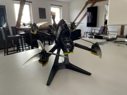 Schwarze FPV-Renn-Drohne mit vier Propellern und sichtbarer Frontkamera auf einem kleinen Ständer auf einem weißen Tisch in einem hellen Wohnraum mit Fenstern und Stühlen im Hintergrund.
