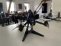 Schwarze FPV-Renn-Drohne mit vier Propellern und sichtbarer Frontkamera auf einem kleinen Ständer auf einem weißen Tisch in einem hellen Wohnraum mit Fenstern und Stühlen im Hintergrund.