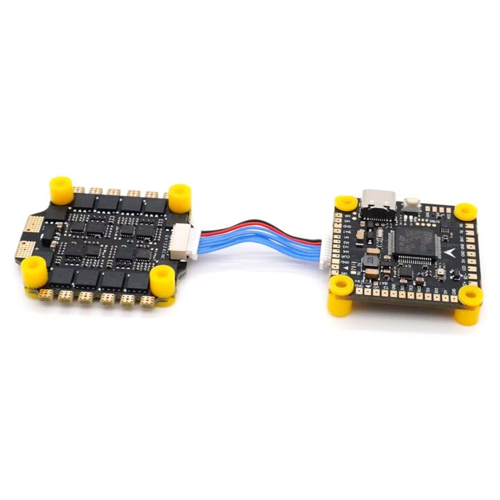 Zwei schwarze FPV-Drohnenplatinen (Flight Controller und ESC) mit gelben Gummistopfen, goldenen Lötpads und einer blauen/roten Flachkabelverbindung; rechts ein Board mit Micro‑USB‑Anschluss auf weißem Hintergrund.