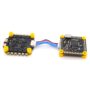 Zwei schwarze FPV-Drohnenplatinen (Flight Controller und ESC) mit gelben Gummistopfen, goldenen Lötpads und einer blauen/roten Flachkabelverbindung; rechts ein Board mit Micro‑USB‑Anschluss auf weißem Hintergrund.