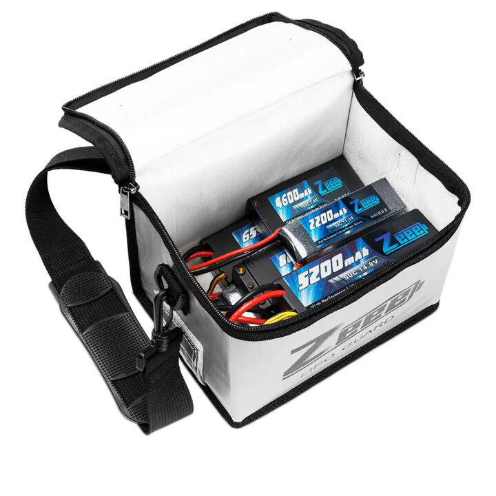 fpv-akku-tasche-sicherer-transport-und-lagerung Geöffnete weiße, gepolsterte Reißverschlusstasche mit verstellbarem schwarzem Schultergurt, in der mehrere blau beschriftete LiPo-Akkus (z. B. 5200 mAh, 2200 mAh) mit roten und schwarzen Anschlusskabeln liegen.