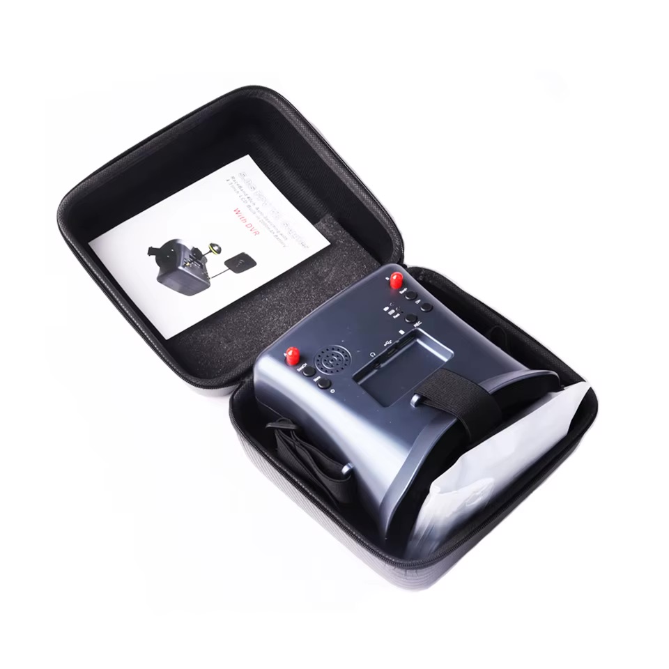 fpv-brille-58g-mit-ladekabel-und-zubehoer FPV‑Drohnen‑Brille in einer offenen schwarzen Tragetasche, dunkelgraue Videobrille mit Kopfband, Bedienelementen und eingebautem Lautsprecher sowie einer Bedienungsanleitung im Deckel.