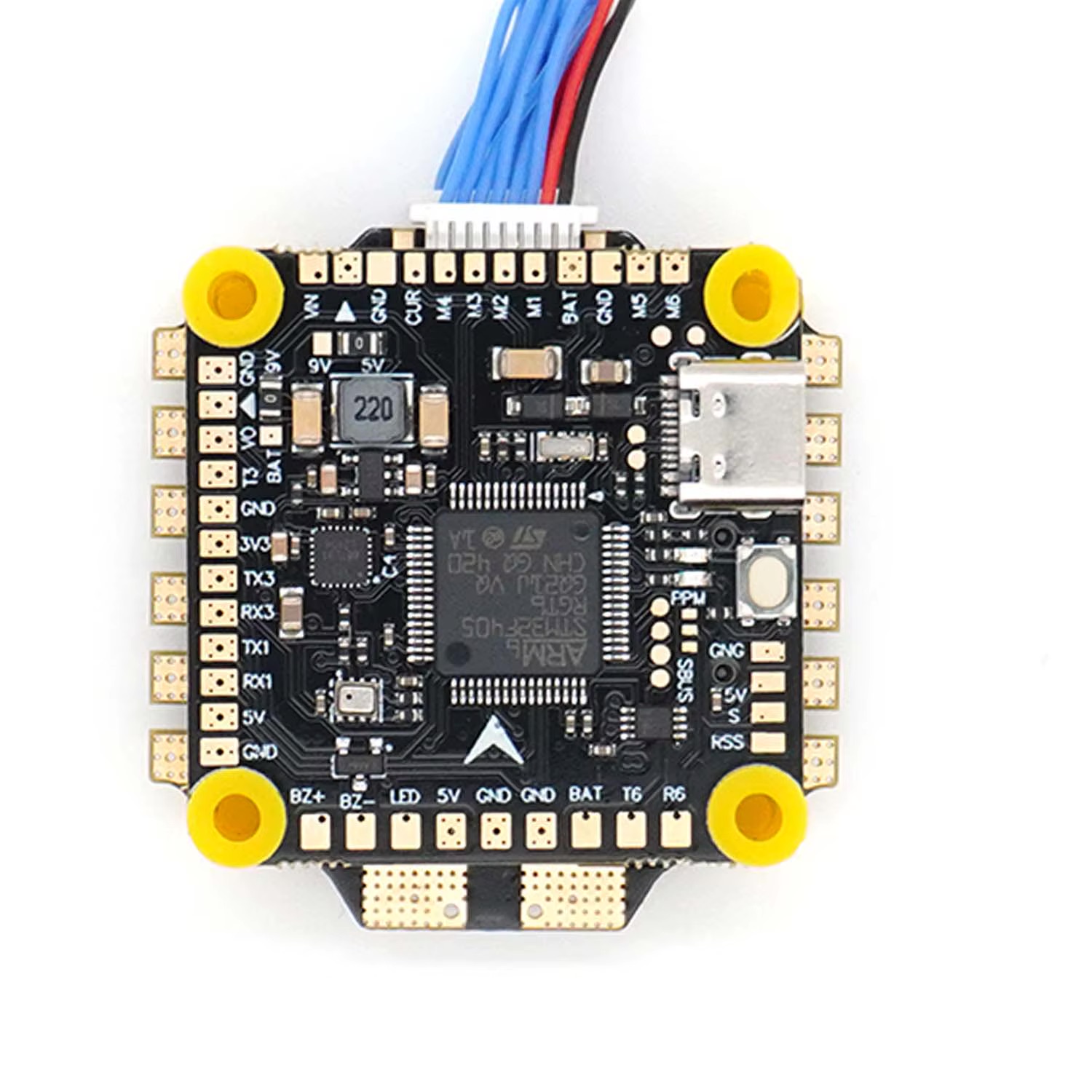 Schwarze Flight‑Controller‑Leiterplatte für FPV‑Drohnen mit zentralem Mikrocontrollerchip, USB‑C‑Anschluss oben rechts, vier gelben Gummitüllen an den Befestigungsbohrungen, zahlreichen goldfarbenen Lötpads und einem mehradrigen Anschlusskabel oben.