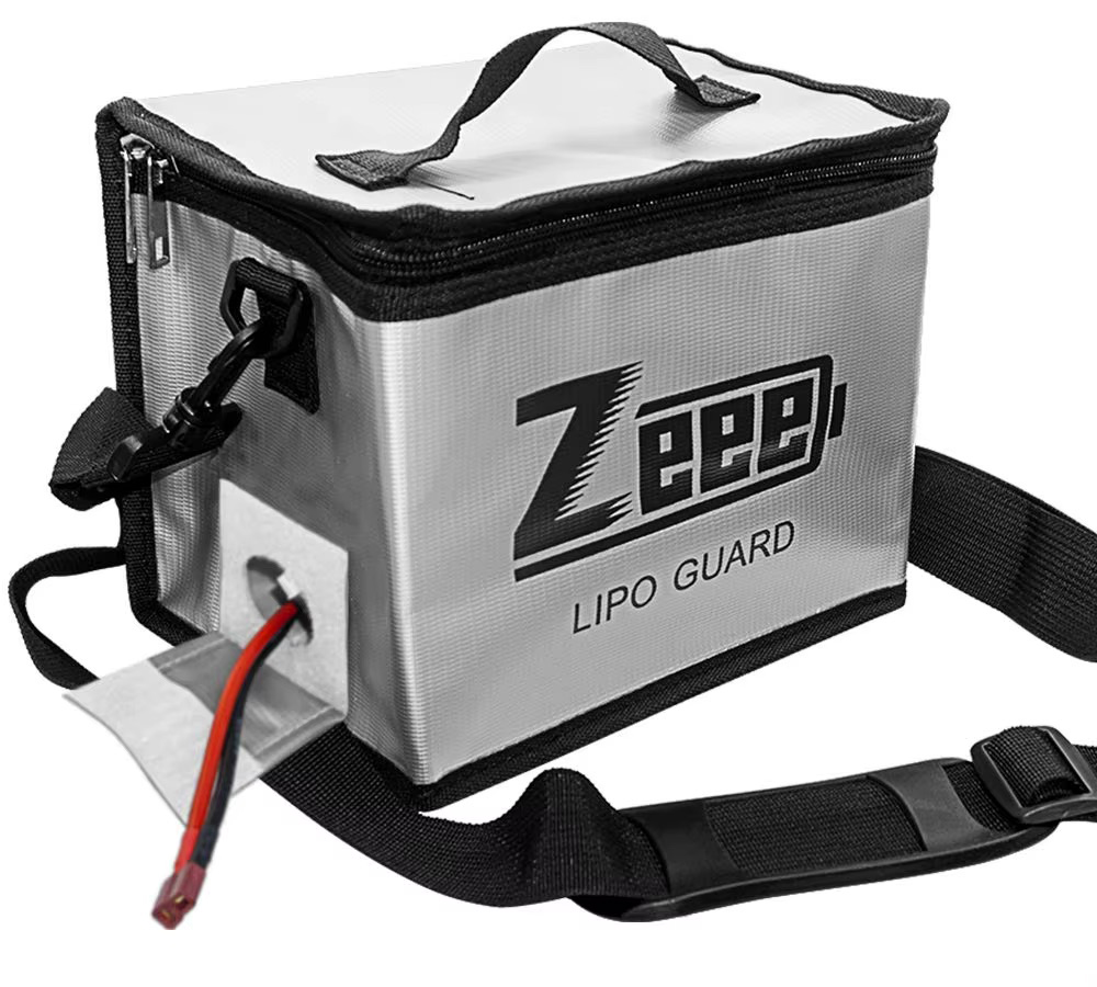 Silberne rechteckige LiPo-Sicherheitsbox mit Reißverschluss, Tragegriff und abnehmbarem Schultergurt, Vorderseite mit Schriftzug „Zeep LIPO GUARD“ und ein rot-schwarzes Batteriekabel, das durch eine seitliche Öffnung führt.