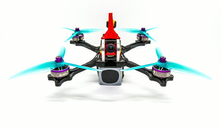 fpv-propeller-gemfan-51466-v2-5-zoll-dreiblatt FPV‑Racing‑Drohne mit vier türkisblauen 5‑Zoll‑Dreiblatt‑Propellern, roter Haube, nach vorn gerichteter Kamera und schwarzem Carbon‑Rahmen vor weißem Hintergrund.