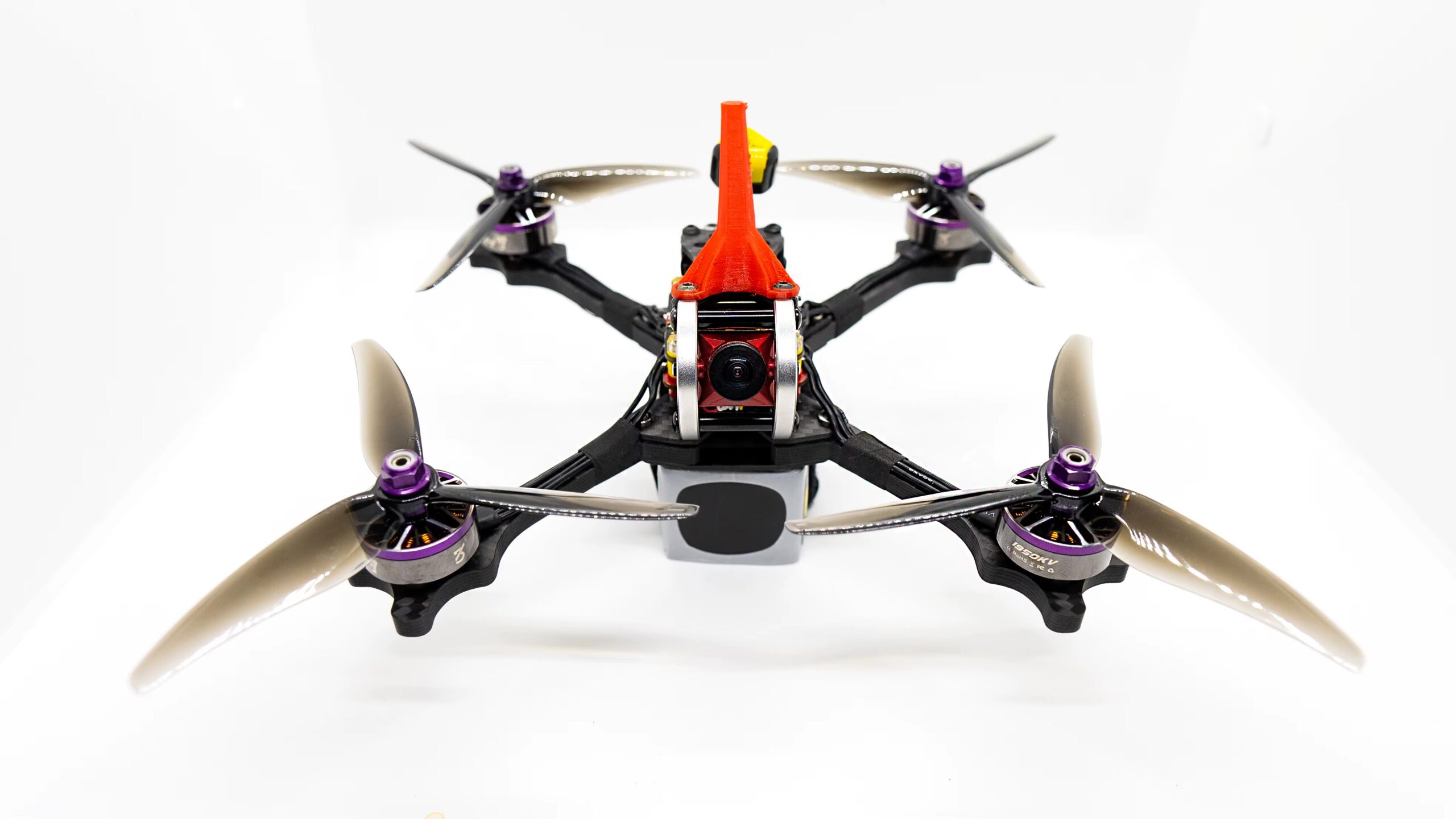 fpv-racing-drone-propeller-gemfan-51466 Frontansicht einer FPV-Renn-Drohne auf weißem Hintergrund mit roter Cockpit-Verkleidung, sichtbarer Kamera, vier Carbon-Armen und vier 5-Zoll-Dreiblatt-Propellern mit lila Motorhauben.