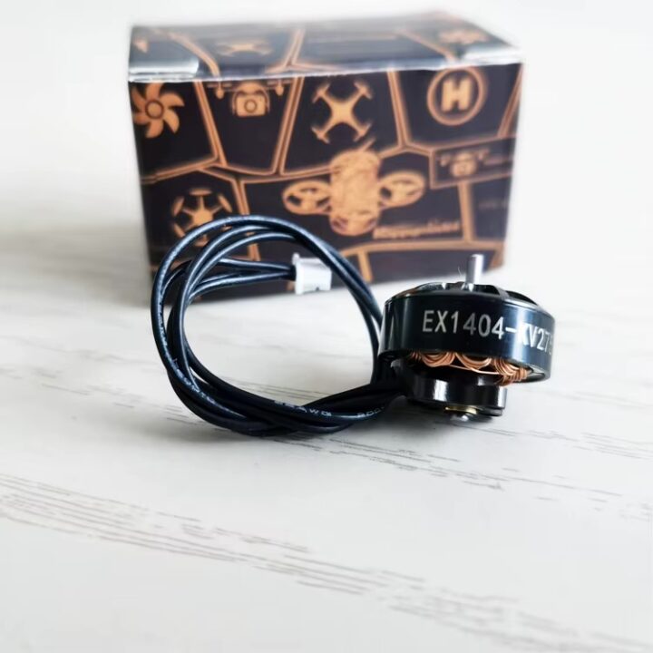 Schwarz glänzender FPV-Drohnen-Brushless-Motor mit sichtbarer Aufschrift EX1404, aufgewickeltem Anschlusskabel und im Vordergrund vor einer schwarz-orangen Verpackung auf hellem Untergrund.