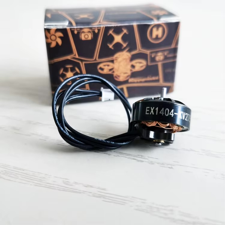 Schwarz glänzender FPV-Drohnen-Brushless-Motor mit sichtbarer Aufschrift EX1404, aufgewickeltem Anschlusskabel und im Vordergrund vor einer schwarz-orangen Verpackung auf hellem Untergrund.
