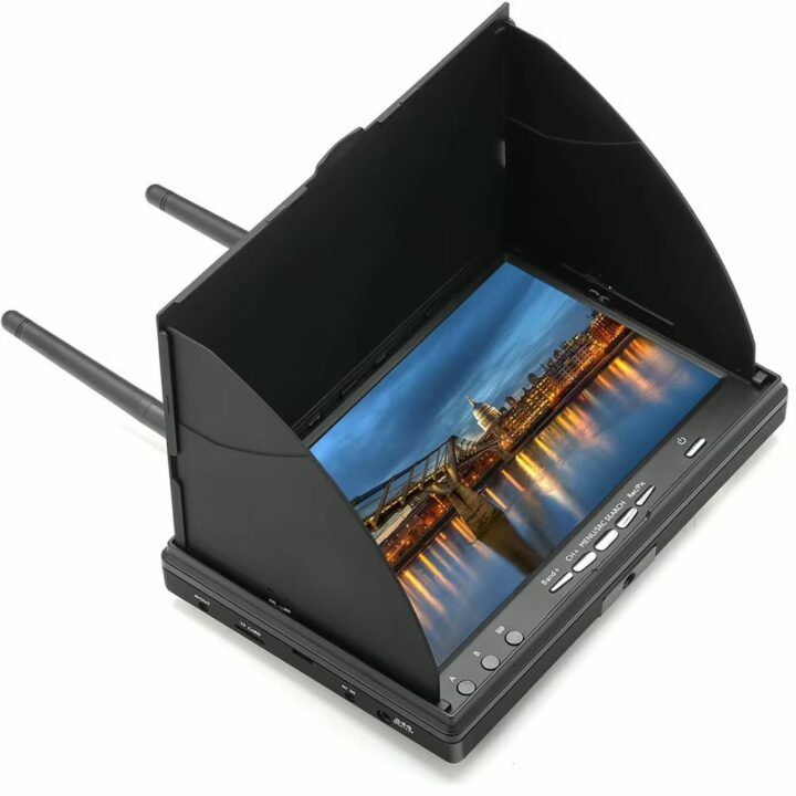 Tragbarer schwarzer FPV-/LCD-Monitor mit aufgesetzter Sonnenblende und zwei ausklappbaren Antennen, zeigt ein abendliches Stadtpanorama mit beleuchteter Brücke und ihrer Spiegelung im Wasser; Bedientasten und Anschlüsse an der Vorderseite sichtbar.