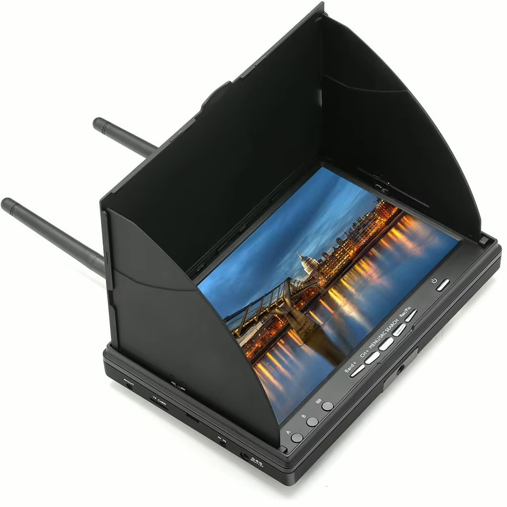 Tragbarer schwarzer FPV-/LCD-Monitor mit aufgesetzter Sonnenblende und zwei ausklappbaren Antennen, zeigt ein abendliches Stadtpanorama mit beleuchteter Brücke und ihrer Spiegelung im Wasser; Bedientasten und Anschlüsse an der Vorderseite sichtbar.