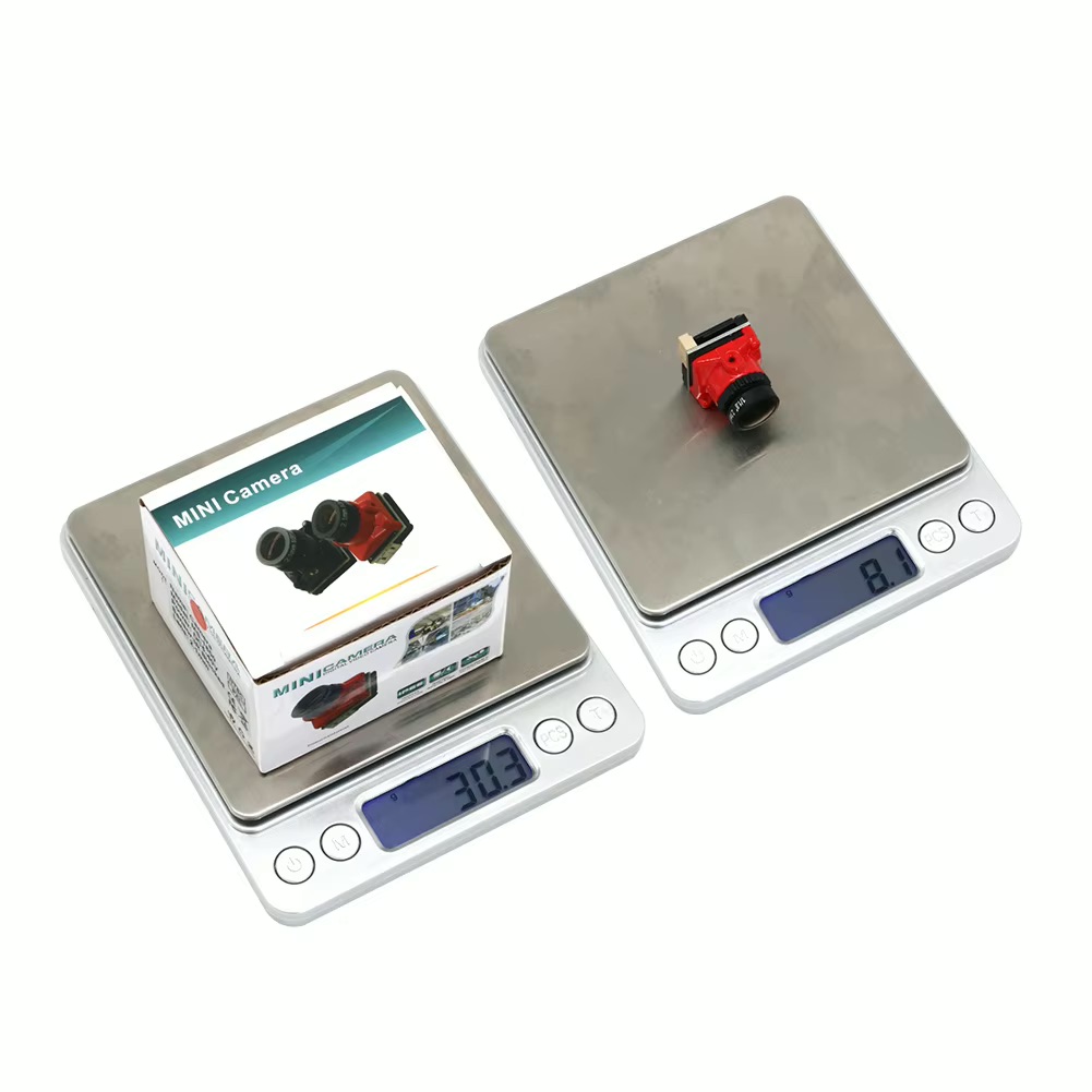 Zwei digitale Waagen mit Edelstahlwiegeflächen: links steht eine verpackte 'MINI Camera' auf der Waage (Display 30,3 g), rechts liegt eine kleine rote Kamerabaugruppe auf der Waage (Display 8,1 g), beide mit blau hintergrundbeleuchteten LCD-Anzeigen.